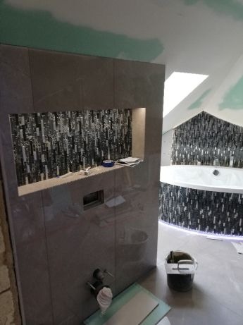 Łazienka w trakcie wykańczania: szara płytka ścienna z wnęką wyłożoną mozaiką, częściowo zabudowana wanna z mozaiką, widoczne narzędzia glazurnicze, ściany częściowo w stanie surowym.