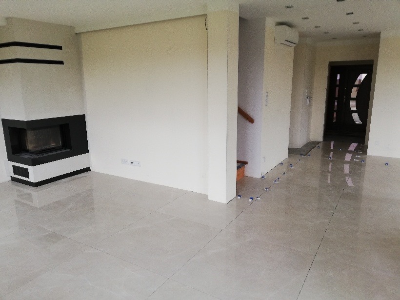 Salon w trakcie układania jasnych, błyszczących płytek podłogowych; widoczne krzyżyki dystansowe, nowoczesny kominek z czarnymi akcentami i wejście do kolejnego pomieszczenia.