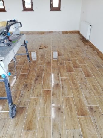 Ułożona podłoga z płytek imitujących drewno w jasnym pomieszczeniu, widoczna przecinarka do płytek i fragment grzejnika.