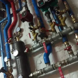 Skomplikowana instalacja hydrauliczna i grzewcza z rurami w izolacji czerwonej i niebieskiej, manometry i zawory na tle ściany z cegły pomalowanej na biało.