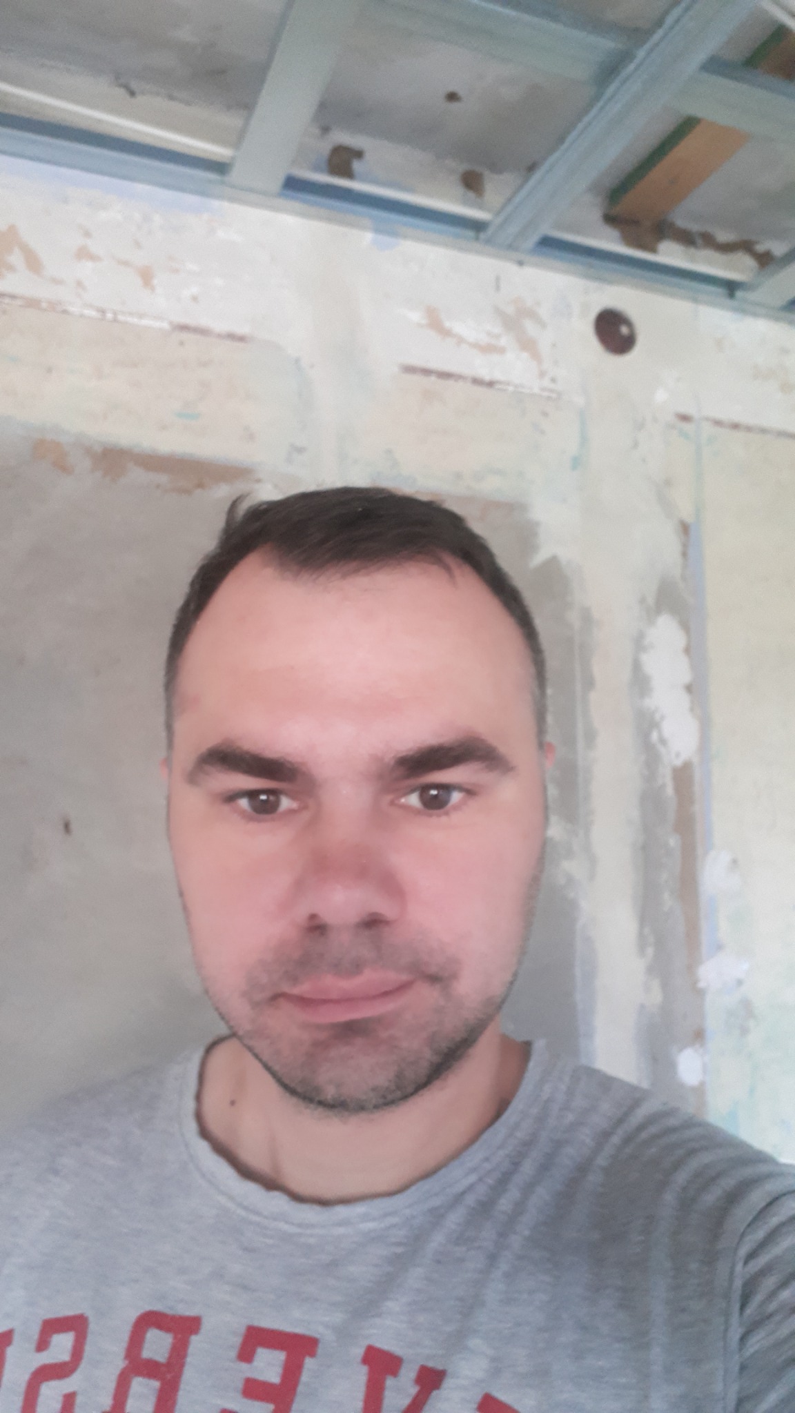 Portret mężczyzny na tle surowej ściany z widocznymi śladami po pracach remontowych i elementami konstrukcji sufitu podwieszanego.