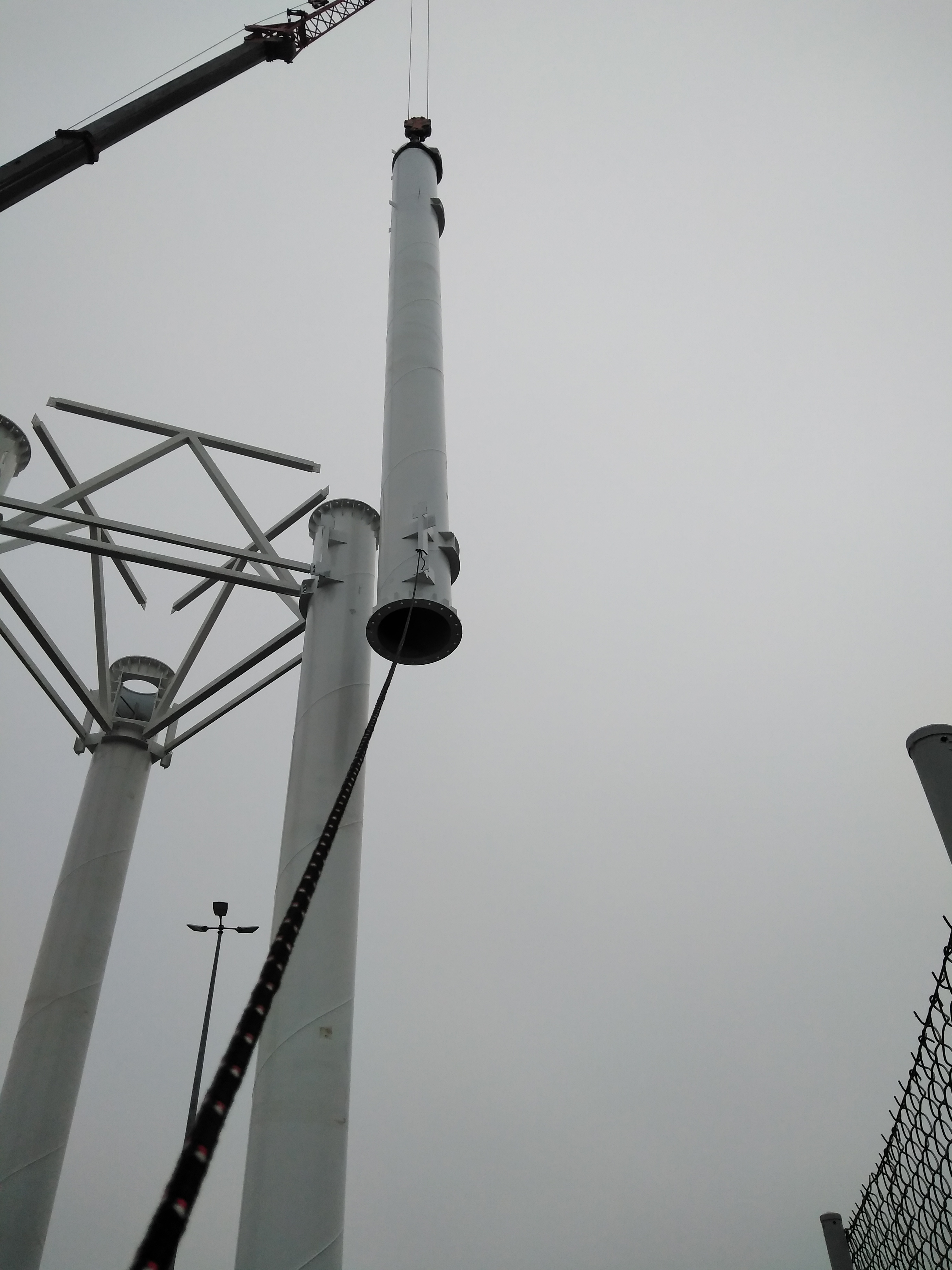 Montaż stalowej kolumny z użyciem dźwigu na tle pochmurnego nieba, widoczne elementy konstrukcji hali w trakcie wznoszenia.