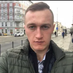 Autoportret mężczyzny w kurtce na tle miejskiej ulicy z budynkami o klasycznej architekturze i zaparkowanymi samochodami.