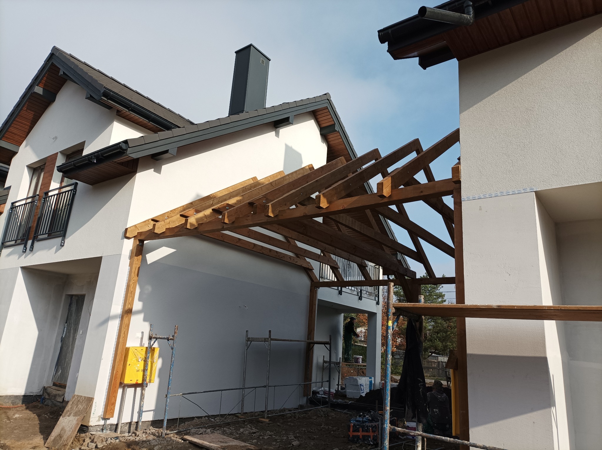 Surowa konstrukcja drewniana zadaszenia łączącego dwa budynki w stanie surowym, widoczne rusztowania i instalacja gazowa na elewacji.