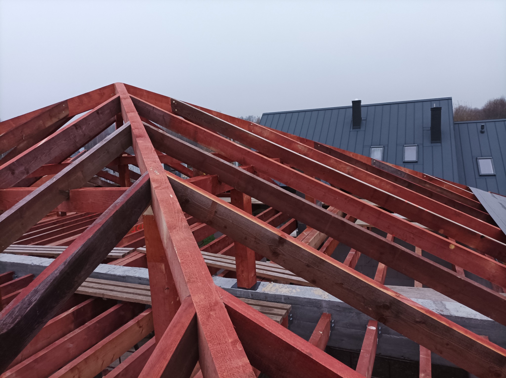 Konstrukcja drewniana dachu domu szkieletowego w trakcie budowy, widoczne czerwono-brązowe belki i elementy mocujące, na tle szarego nieba i fragmentu dachu sąsiedniego budynku.