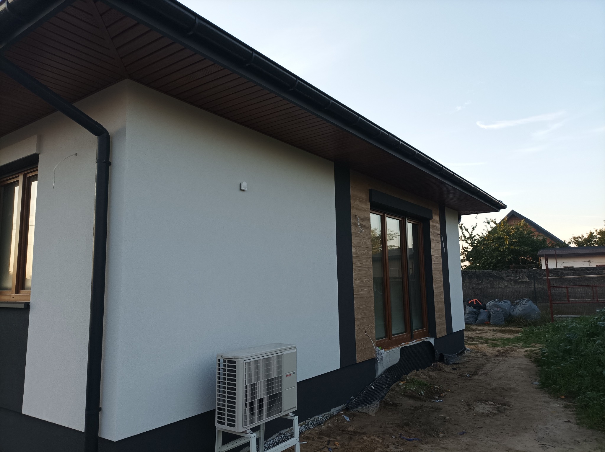 Fragment elewacji budynku z jasnym tynkiem, drewnopodobnymi elementami dekoracyjnymi, klimatyzatorem na zewnątrz i czarną rynną.