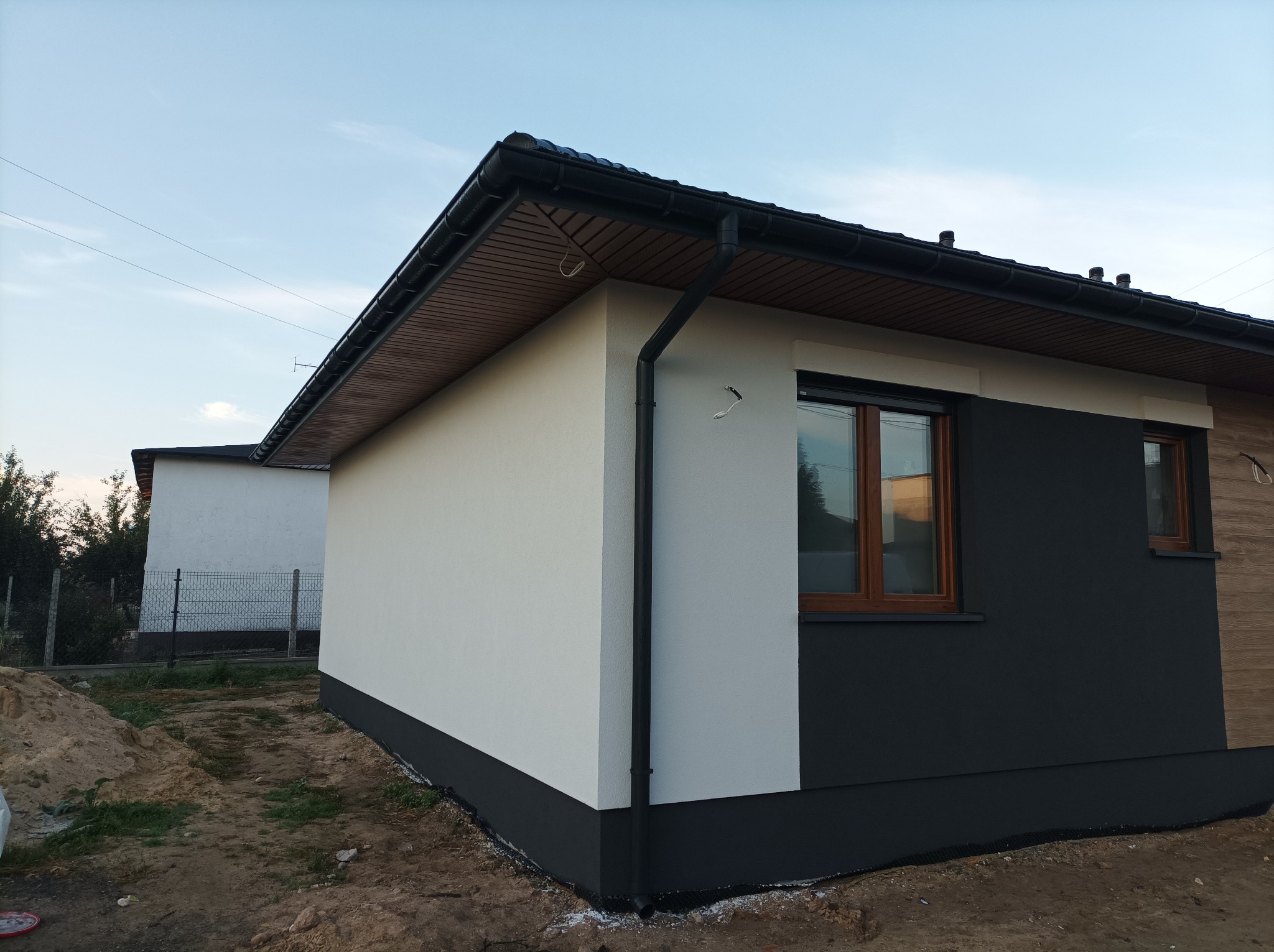 Wykończony zewnętrznie dom szkieletowy z dwukolorową elewacją (biały tynk i grafitowa farba), widoczne okna z drewnianą stolarką i czarne rynny, teren wokół domu w trakcie zagospodarowywania.