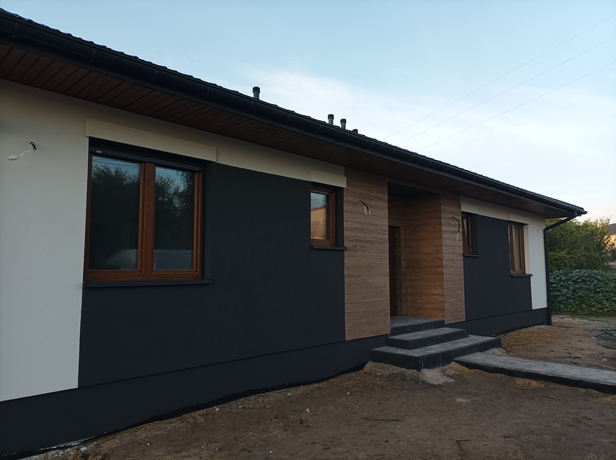 Nowoczesny dom z elewacją w kolorach bieli i grafitu, drewniane okna, wejście wykończone drewnopodobną okładziną, schody z kostki brukowej, teren wokół domu w trakcie prac wykończeniowych.