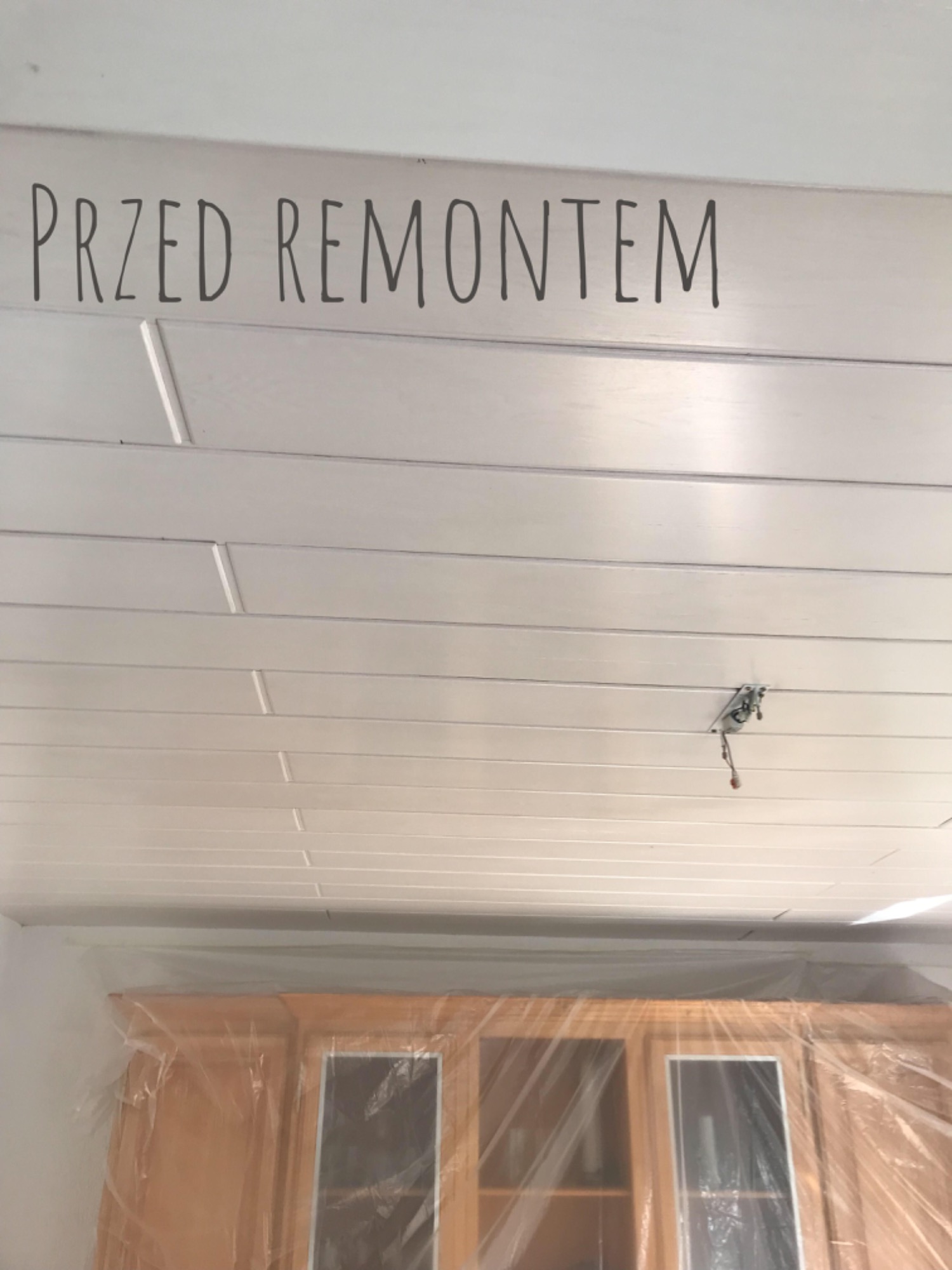 Zdjęcie z Gąsawy przedstawia biały, drewniany sufit panelowy z napisem 'PRZED REMONTEM', a poniżej zabezpieczone folią meble kuchenne, ukazujące stan pomieszczenia przed rozpoczęciem prac.