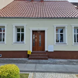 Odnowiona fasada domu jednorodzinnego z nową stolarką okienną i drewnianymi drzwiami wejściowymi, widoczne detale architektoniczne i fragment chodnika z kostki brukowej.