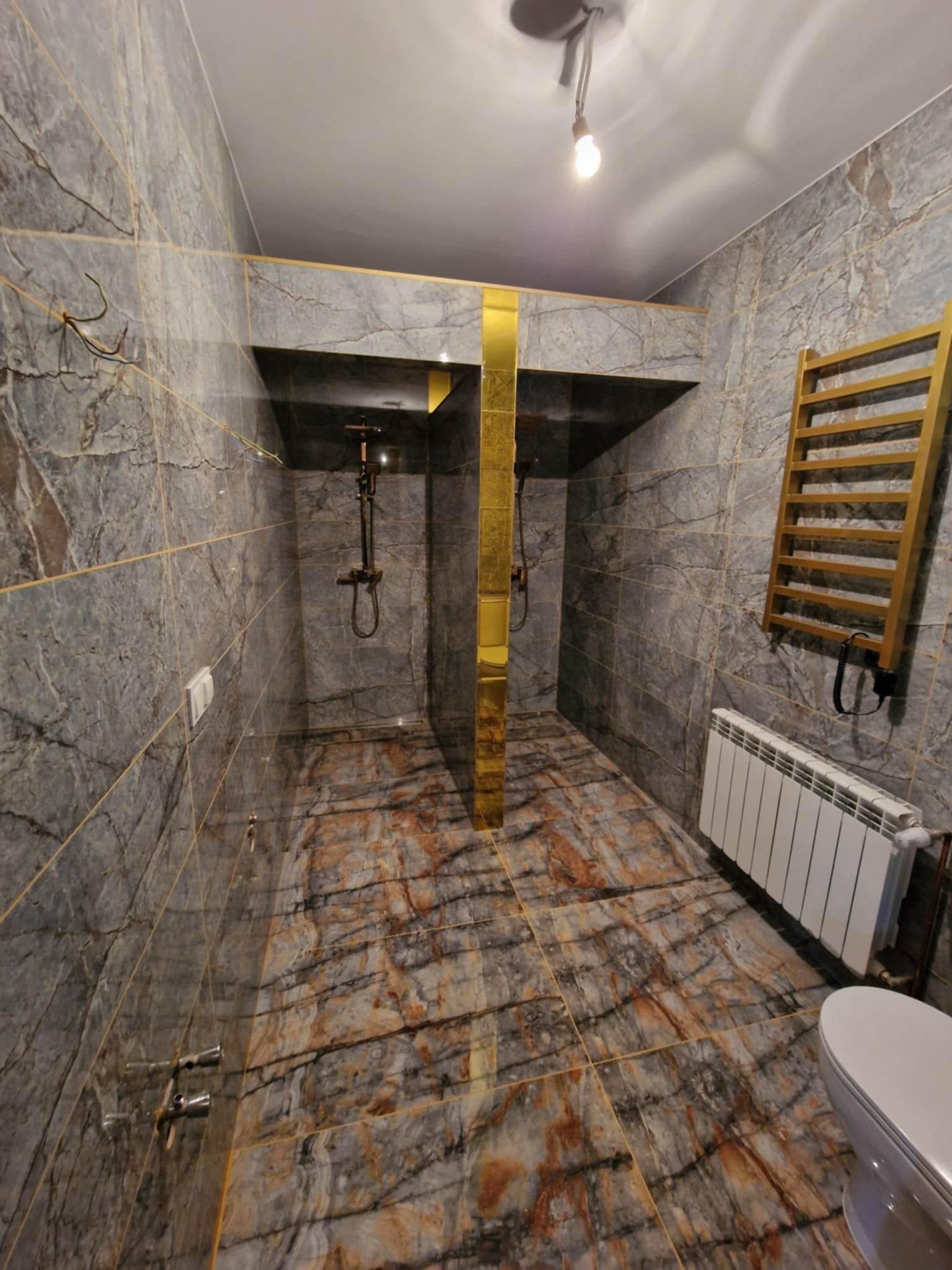 Nowoczesna łazienka z dwoma kabinami prysznicowymi oddzielonymi złotym panelem, marmurowe płytki na ścianach i podłodze, grzejnik drabinkowy w kolorze złota oraz biały grzejnik panelowy.