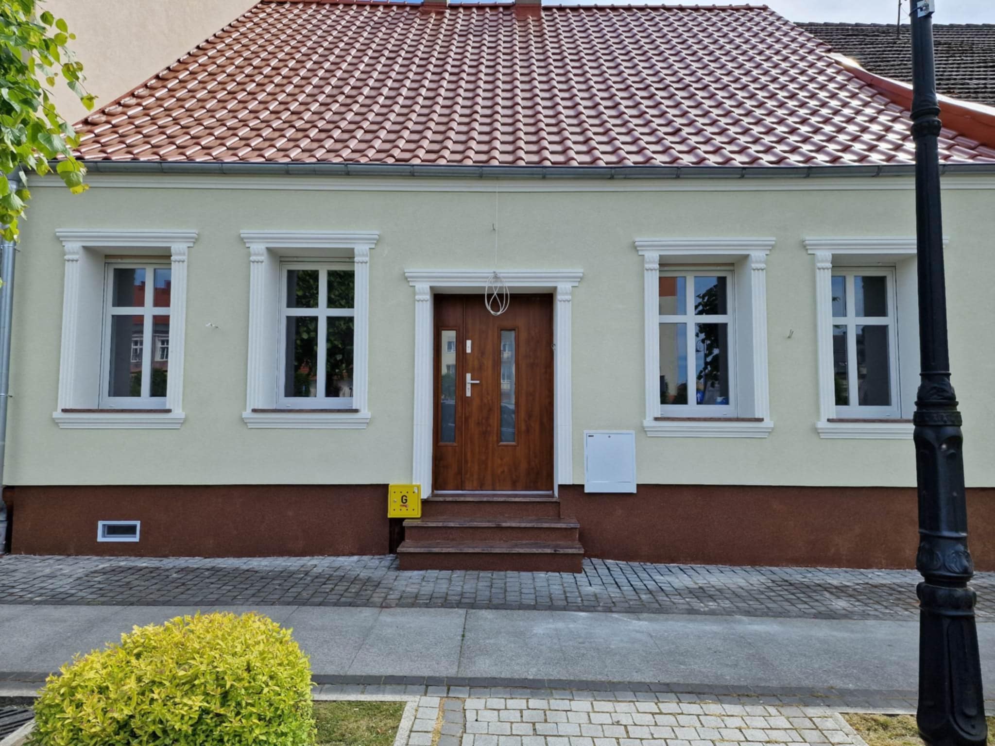 Odnowiona fasada domu jednorodzinnego z nową stolarką okienną i drewnianymi drzwiami wejściowymi, widoczne detale architektoniczne i fragment chodnika z kostki brukowej.