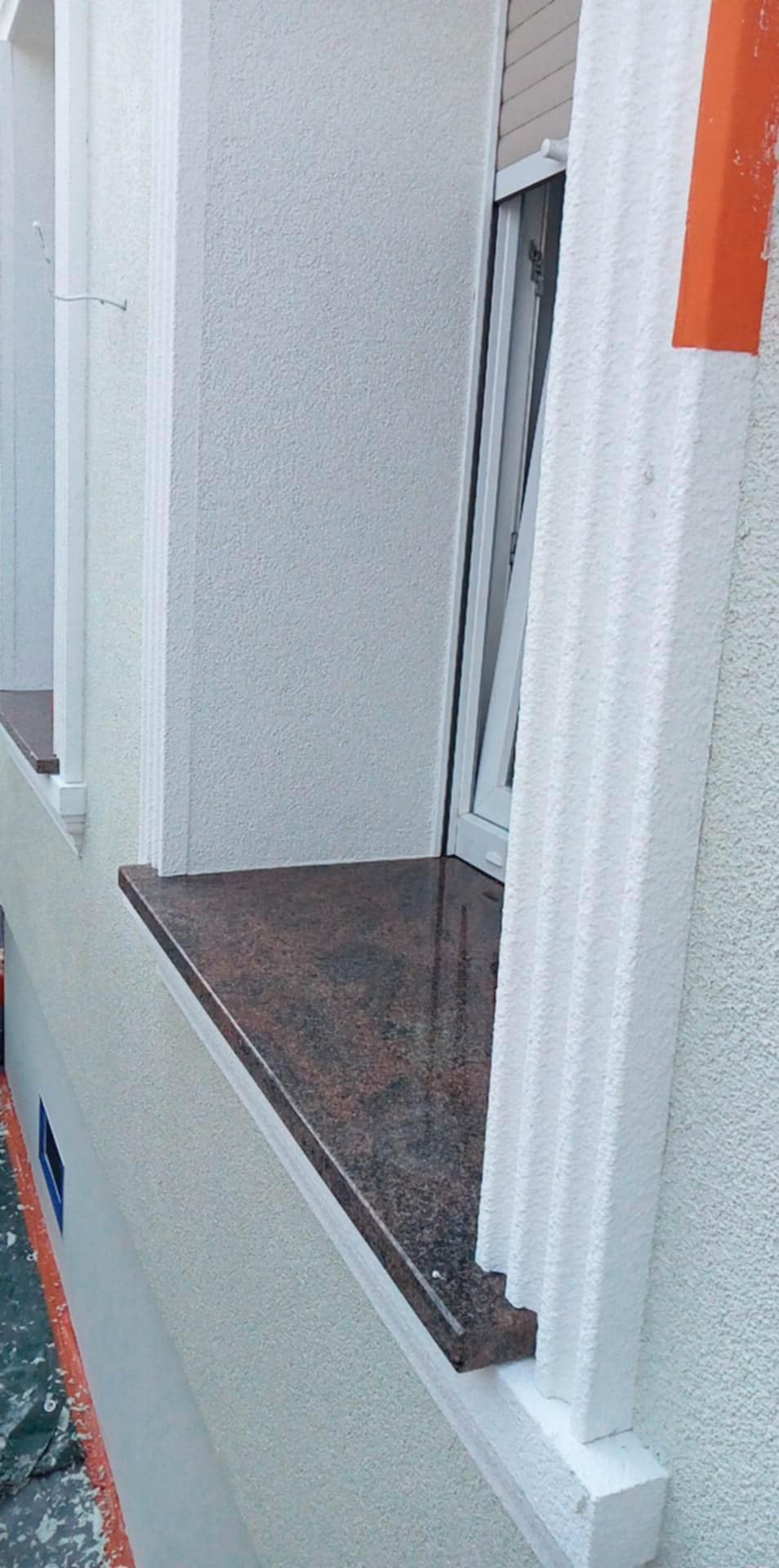 Granitowy parapet zewnętrzny, detal elewacji z oknem, biała struktura tynku z dekoracyjnymi pilastrami.