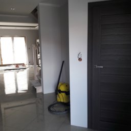 Krzysztof Błauciak - Wnętrze w trakcie wykańczania: szare płytki podłogowe, fragment drewnianych schodów, żółty odkurzacz przemysłowy, szara, panelowa  ościeżnica i drzwi, wystające kable ze ściany.