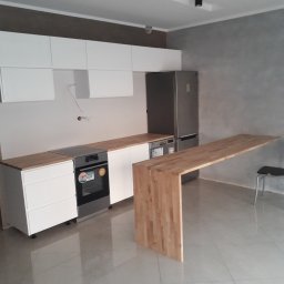 Krzysztof Błauciak - Nowoczesna kuchnia z białymi szafkami, drewnianym blatem i wyspą, szarą ścianą i błyszczącą podłogą z widocznymi przewodami elektrycznymi na ścianie.