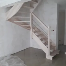 Jasne, drewniane schody z metalowymi barierkami, zamontowane w surowym wnętrzu z widocznymi śladami prac wykończeniowych na ścianach i podłodze.