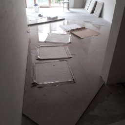 Krzysztof Błauciak - Układanie płytek podłogowych w jasnym pomieszczeniu, zabezpieczone kartonem, widoczny kubeł z narzędziami i okno z widokiem na zieleń.