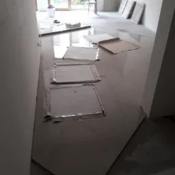 Układanie płytek podłogowych w jasnym pomieszczeniu, zabezpieczone kartonem, widoczny kubeł z narzędziami i okno z widokiem na zieleń.