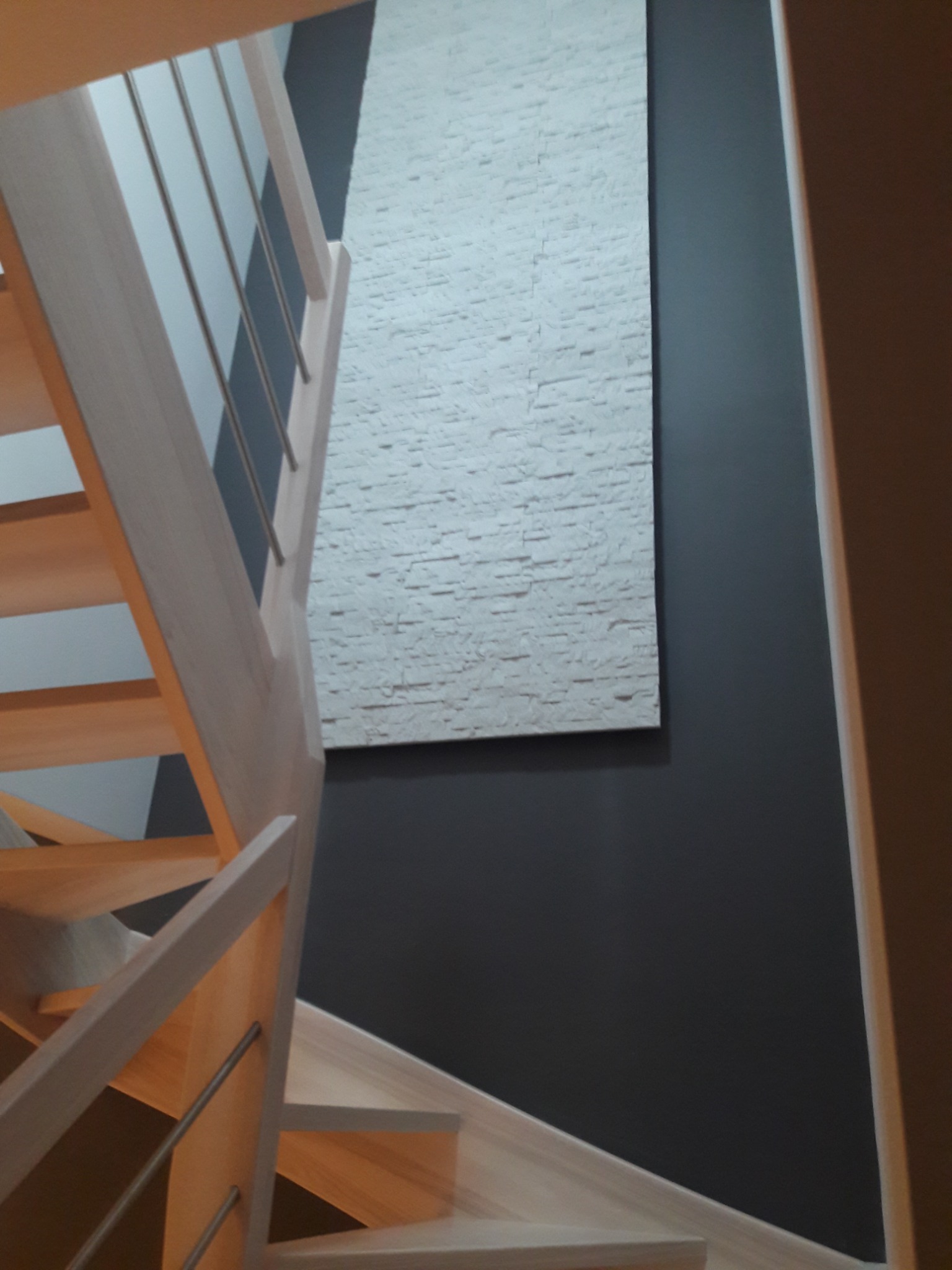 Jasne drewniane schody z metalowymi balustradami, widok z góry, na ścianie dekoracyjna biała okładzina imitująca cegłę na ciemnoszarym tle