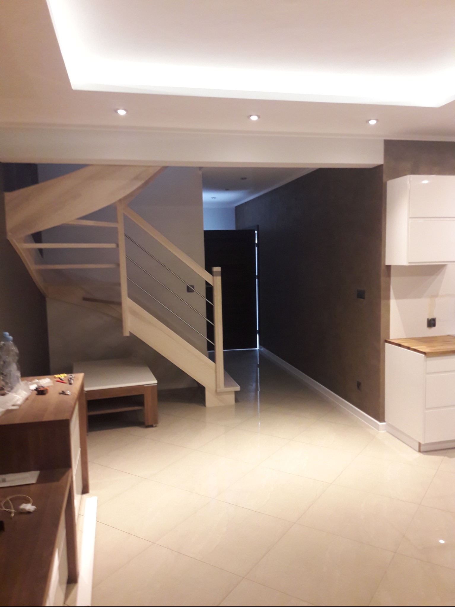 Jasne drewniane schody ze stalowymi relingami w nowoczesnym wnętrzu z beżową podłogą i widoczną częścią białej zabudowy kuchennej.