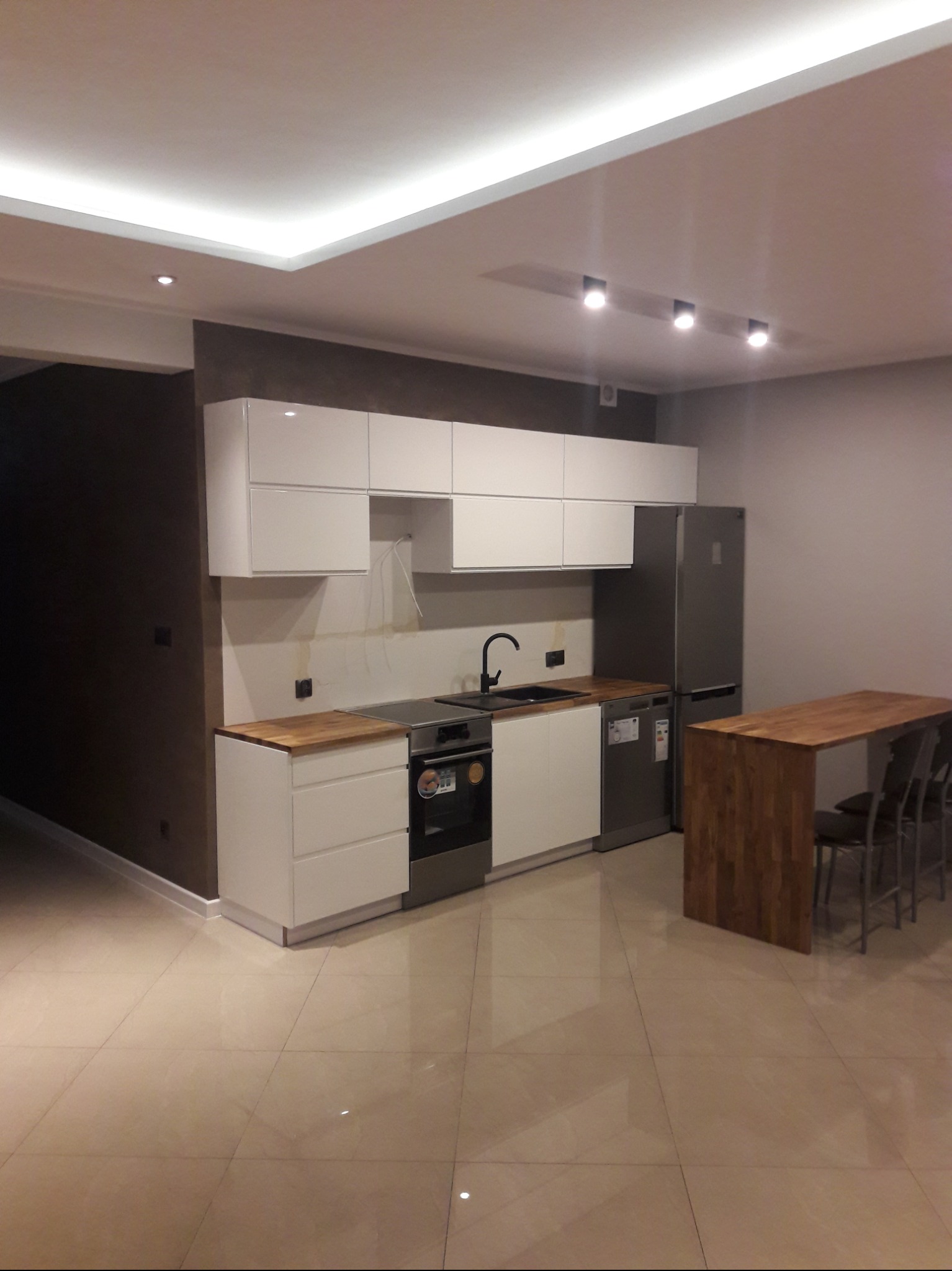 Nowoczesna, jasna kuchnia z białymi szafkami, drewnianymi blatami i stołem barowym z krzesłami, oświetlona ledowym paskiem w suficie i punktowymi lampami.