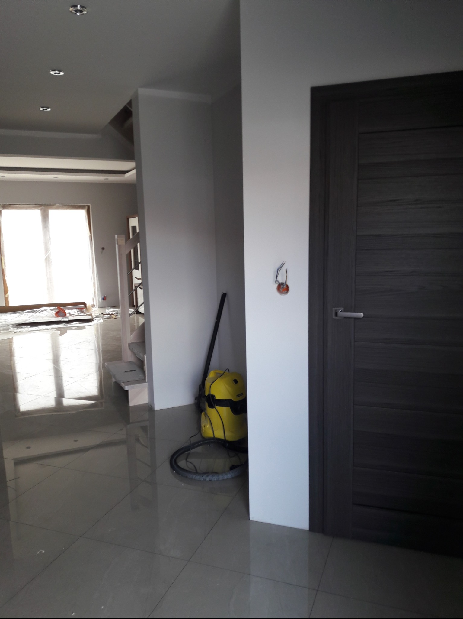 Wnętrze w trakcie wykańczania: szare płytki podłogowe, fragment drewnianych schodów, żółty odkurzacz przemysłowy, szara, panelowa  ościeżnica i drzwi, wystające kable ze ściany.