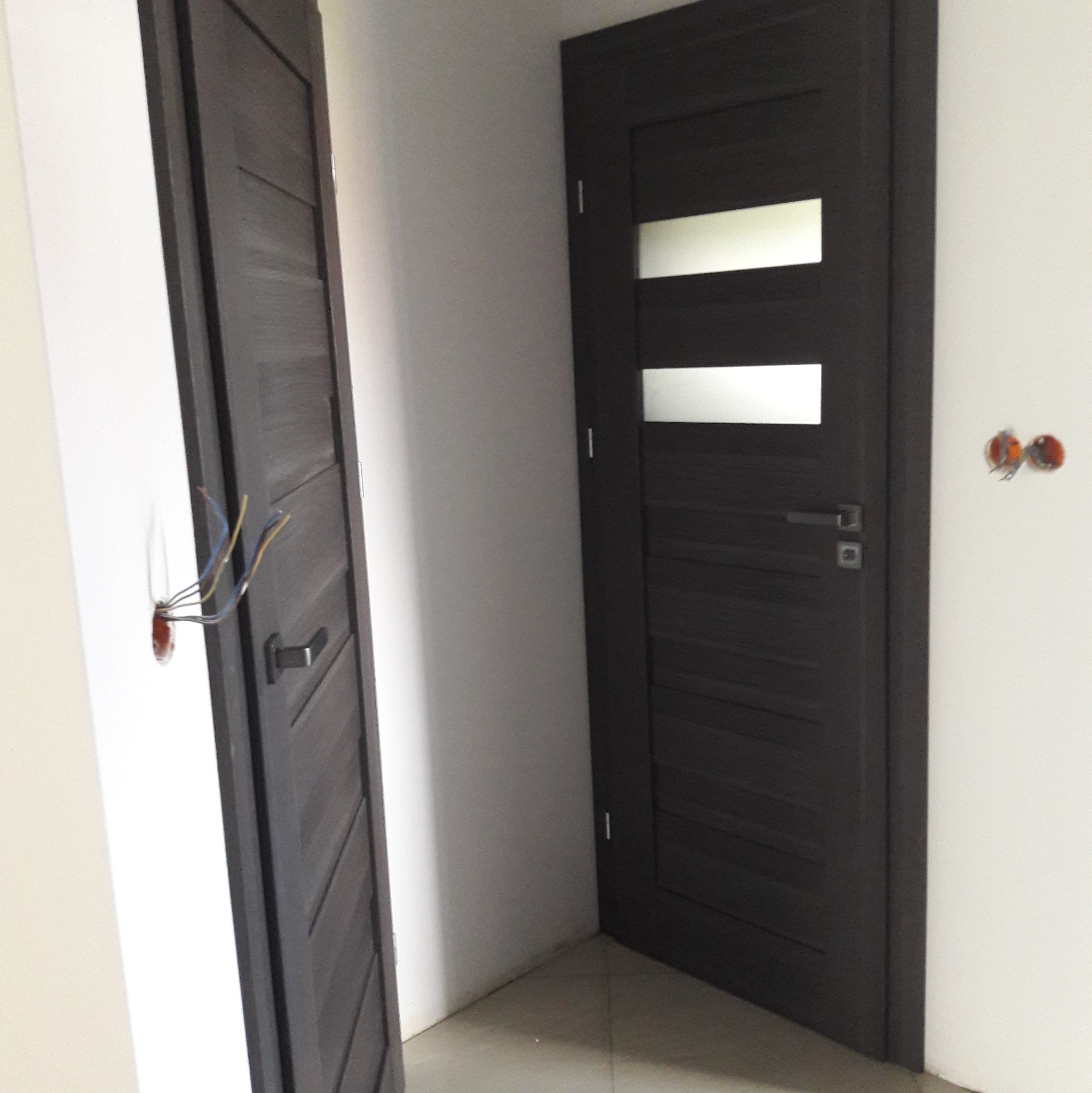 Montaż dwóch ciemnych, drewnopodobnych drzwi wewnętrznych z przeszkleniami w nowym mieszkaniu; widoczne wystające przewody elektryczne obok ościeżnicy.