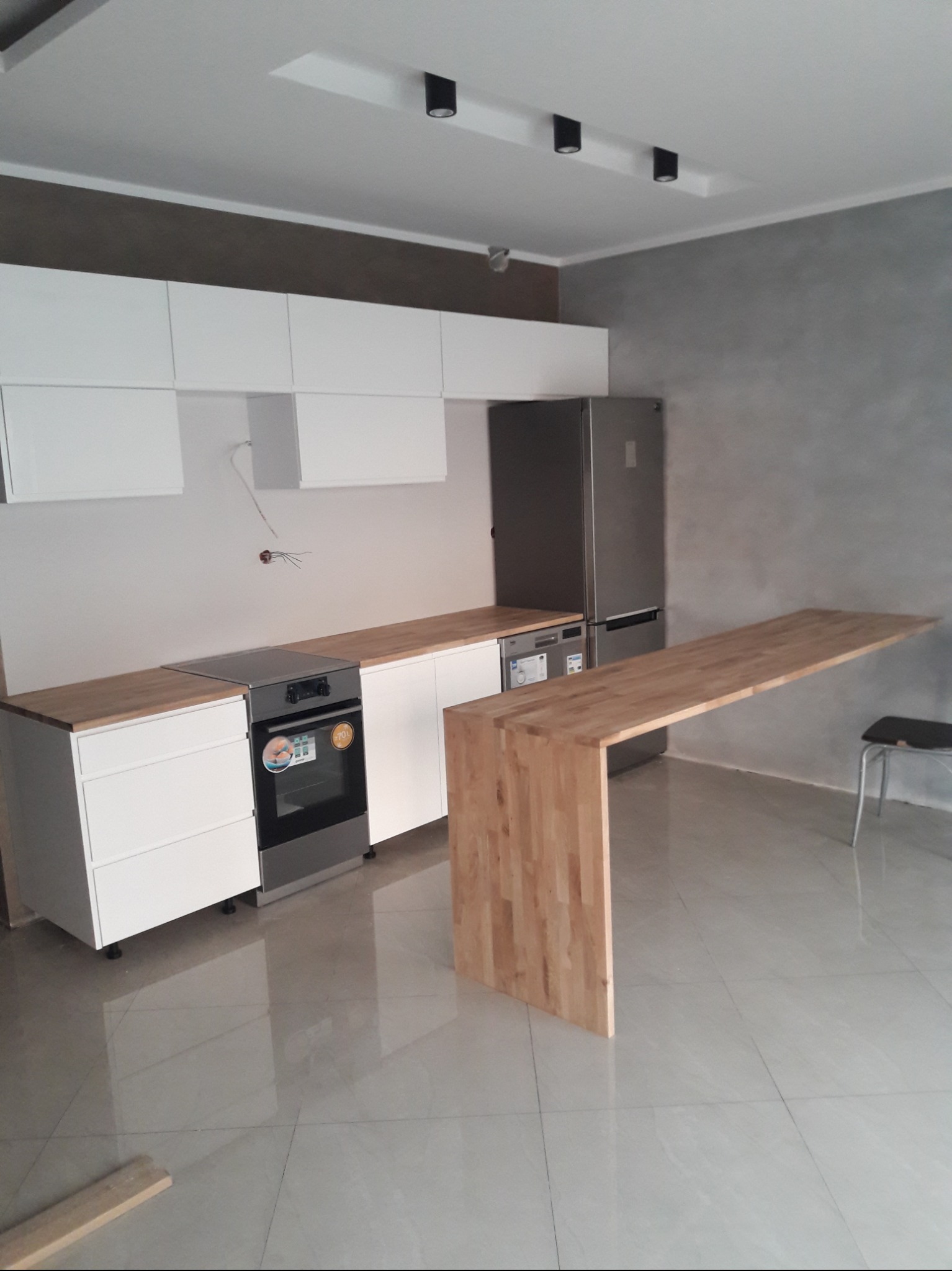 Nowoczesna kuchnia z białymi szafkami, drewnianym blatem i wyspą, szarą ścianą i błyszczącą podłogą z widocznymi przewodami elektrycznymi na ścianie.