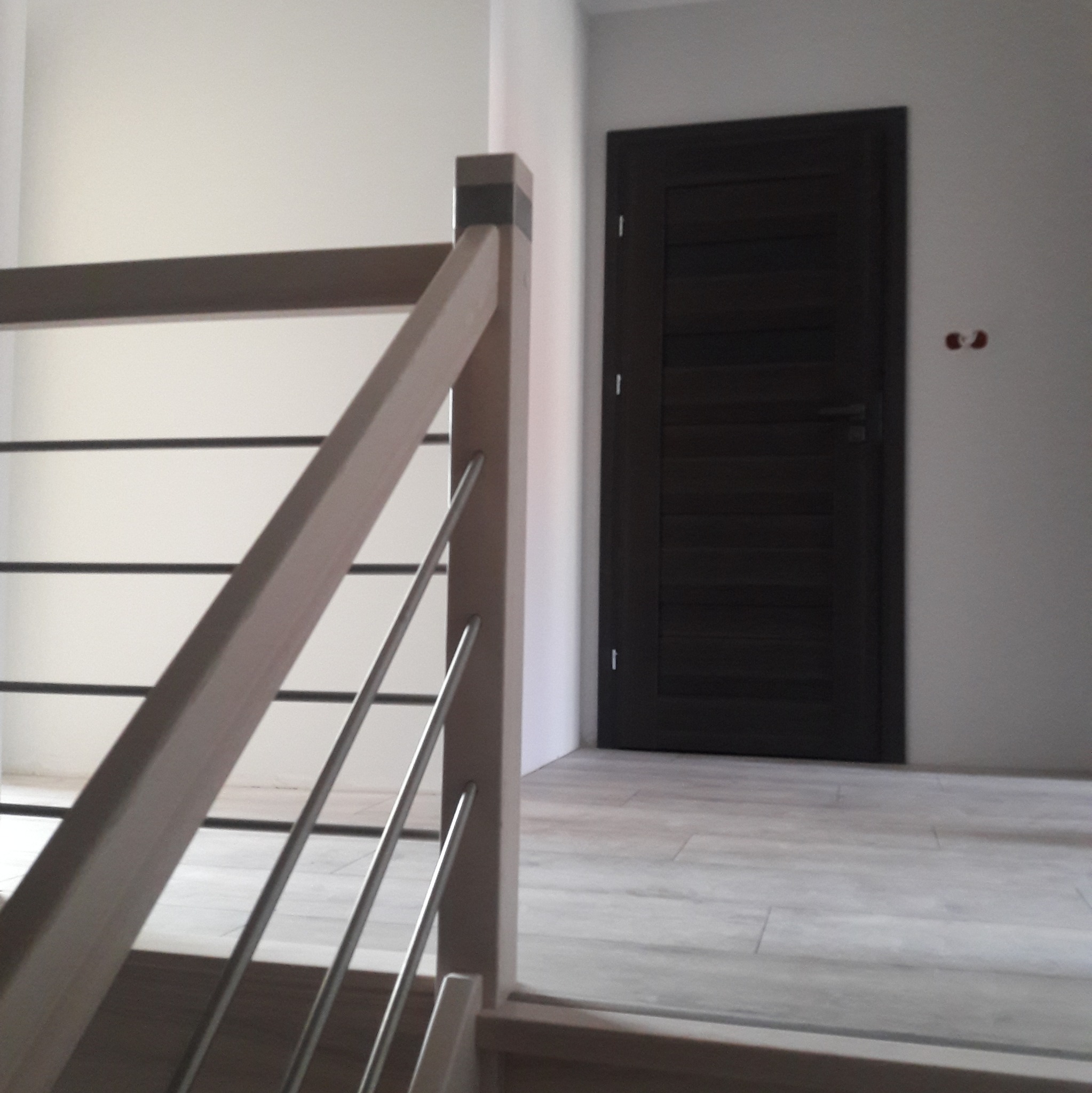 Fragment klatki schodowej z drewnianą balustradą z metalowymi elementami i ciemnymi drzwiami w tle na podłodze z szarych płytek imitujących drewno.