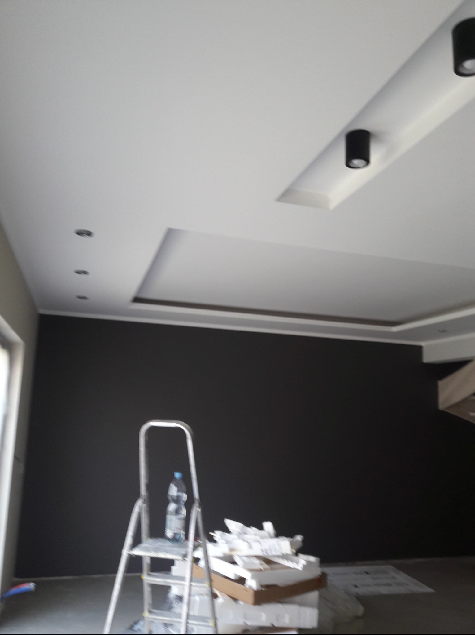 Wnętrze w trakcie prac wykończeniowych: czarna ściana, biały sufit z wbudowanym oświetleniem punktowym, aluminiowa drabina z butelką wody i stos kartonów z materiałami budowlanymi.