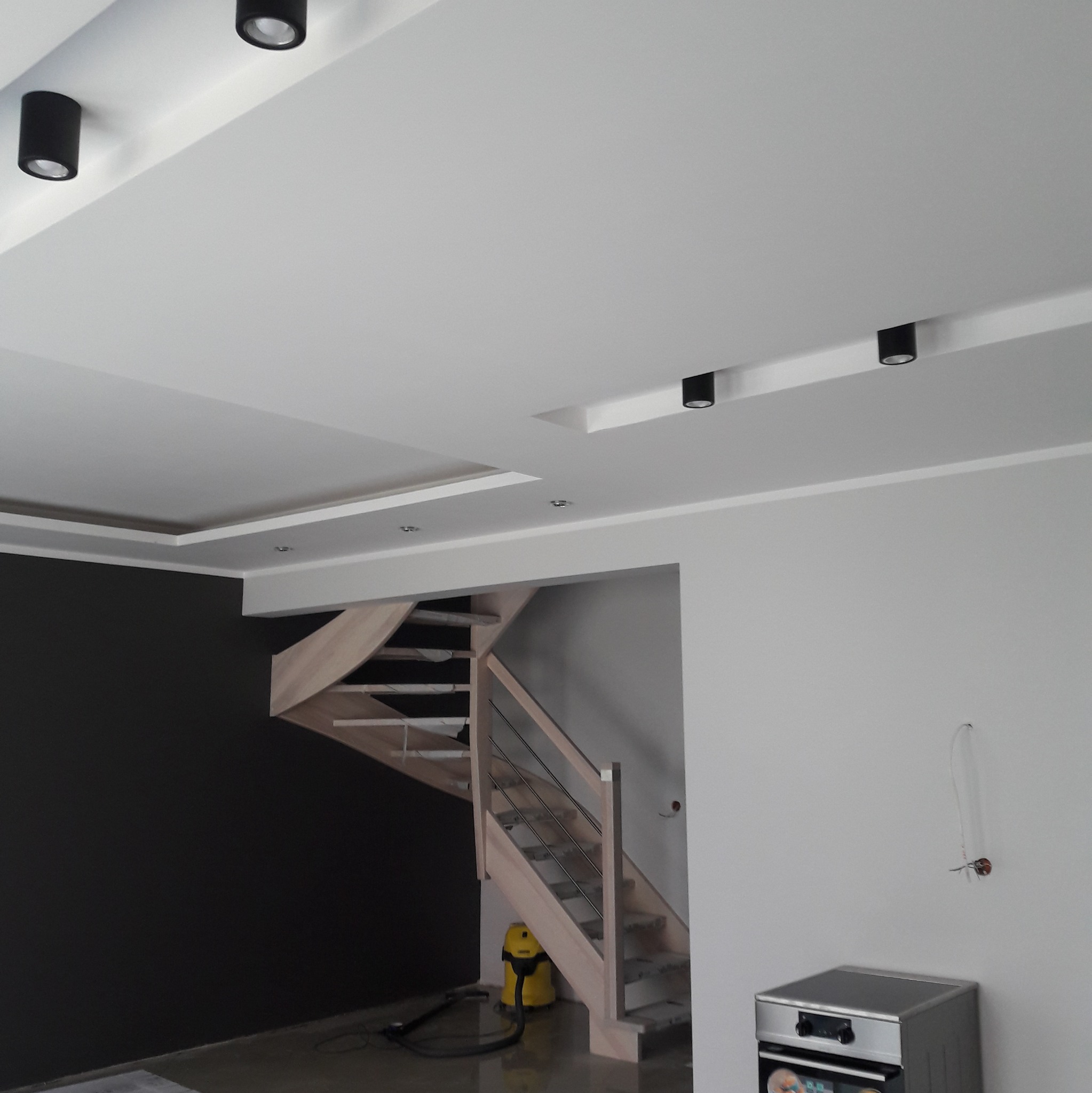 Nowoczesne wnętrze z drewnianymi schodami spiralnymi, czarną ścianą akcentującą i białym sufitem z punktowym oświetleniem, widoczny odkurzacz i niedokończone podłączenie elektryczne.