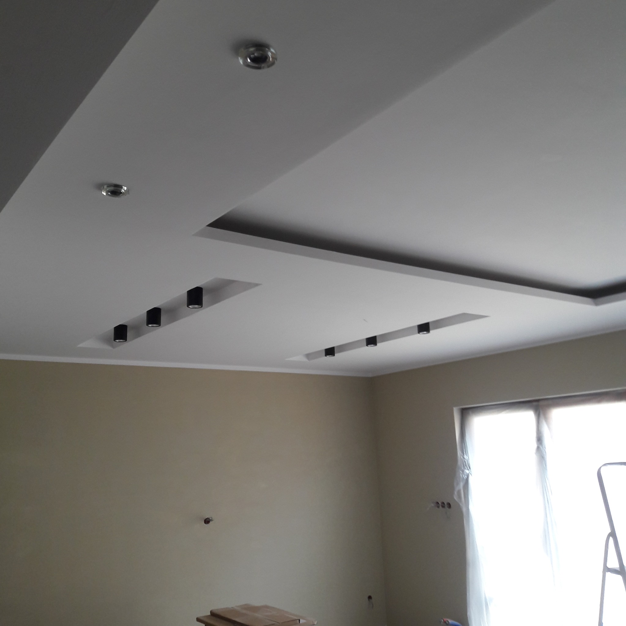 Wykończony sufit podwieszany z oświetleniem punktowym w nowym mieszkaniu, widoczne przygotowane ściany do malowania, okno zabezpieczone folią malarską.