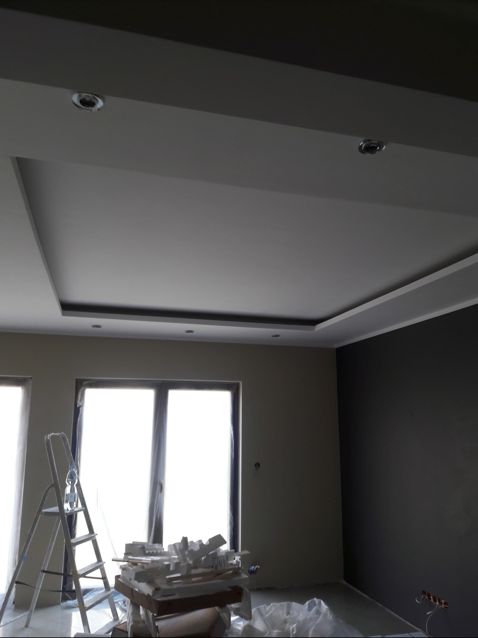 Wnętrze w trakcie wykończenia: widoczny sufit podwieszany z oświetleniem punktowym, drabina oparta o okno zabezpieczone folią, oraz fragment ściany pomalowanej na ciemny kolor.