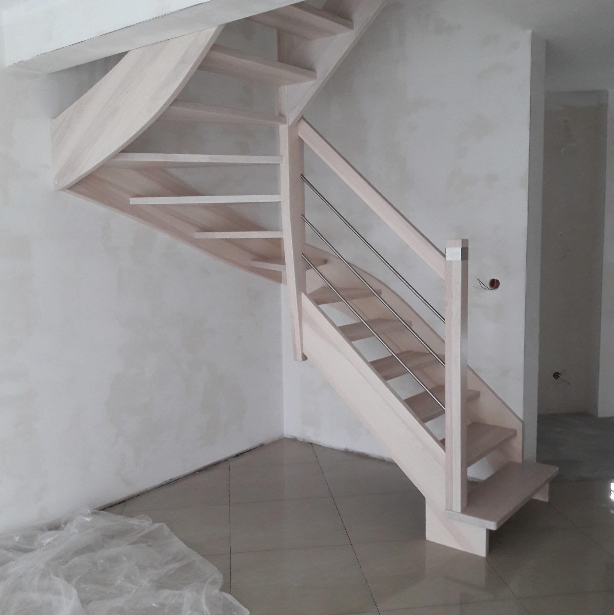 Jasne, drewniane schody z metalowymi barierkami, zamontowane w surowym wnętrzu z widocznymi śladami prac wykończeniowych na ścianach i podłodze.