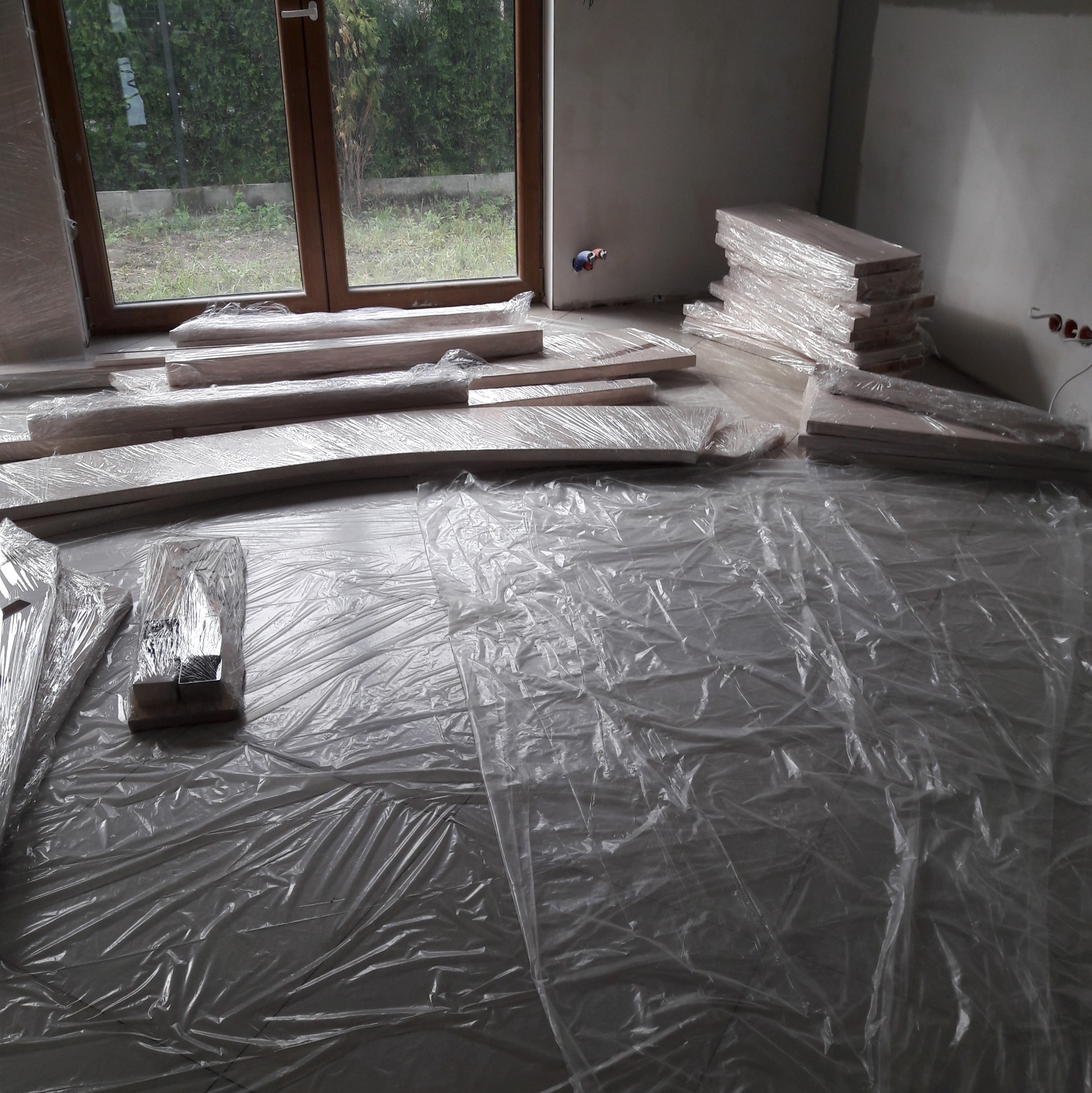 Przygotowanie do ułożenia parkietu: deski podłogowe w folii ochronnej, rozłożone na folii malarskiej w surowym wnętrzu z oknem na ogród.