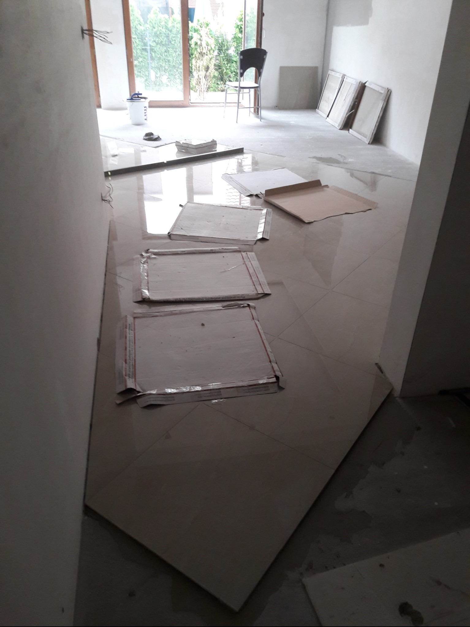 Układanie płytek podłogowych w jasnym pomieszczeniu, zabezpieczone kartonem, widoczny kubeł z narzędziami i okno z widokiem na zieleń.