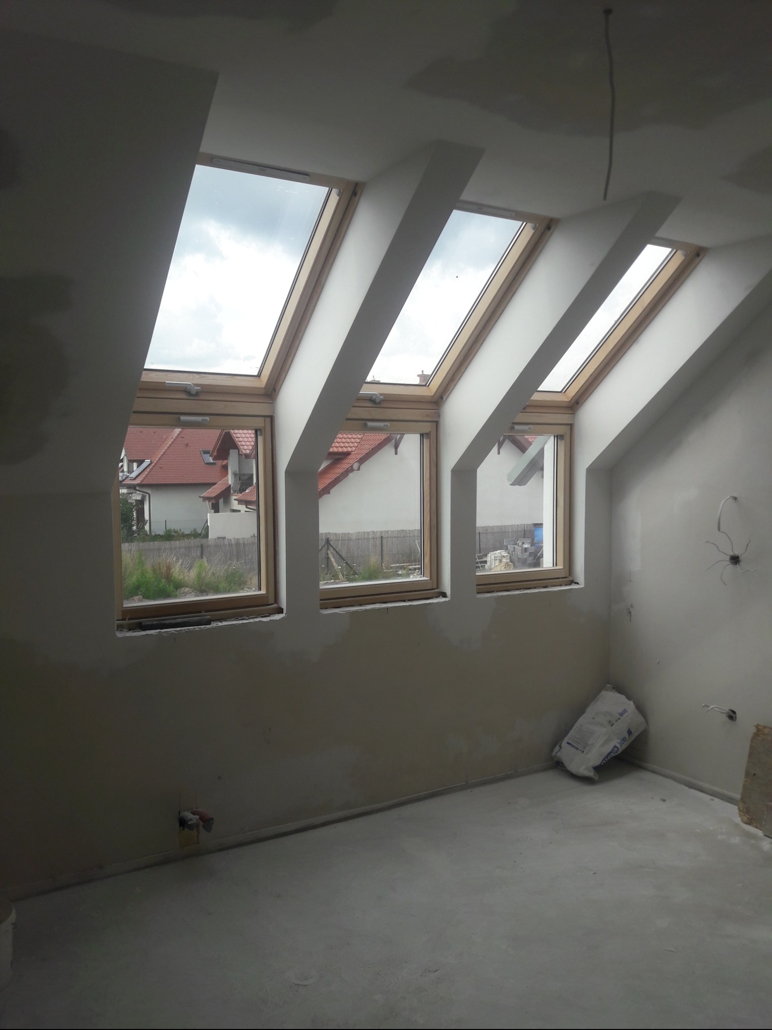 Poddasze w trakcie wykończenia z trzema drewnianymi oknami dachowymi, widok na domy za oknem, ściany w trakcie gładzenia, widoczne przewody elektryczne i worek z zaprawą w rogu.