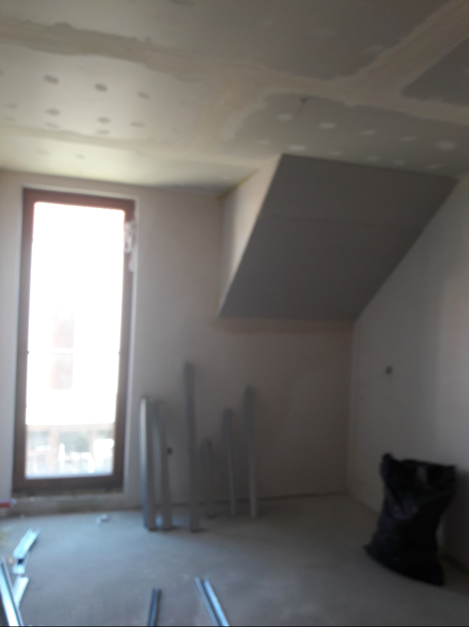 Pokój w trakcie prac wykończeniowych: zamontowana płyta karton gips na skośnym suficie, widoczne profile aluminiowe, okno z widokiem na zewnątrz, czarny worek na śmieci w rogu.