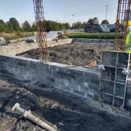 Wykop pod budynek z częściowo wylanymi fundamentami z betonowych bloczków, widoczne stalowe zbrojenia, szalunki, oraz pracownik w żółtym kasku i kamizelce odblaskowej. W tle zaparkowane samochody.