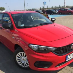 Czerwony samoch&oacute;d osobowy marki Fiat zaparkowany na parkingu z otwartymi drzwiami, widoczne inne pojazdy i znak dla niepełnosprawnych w tle.