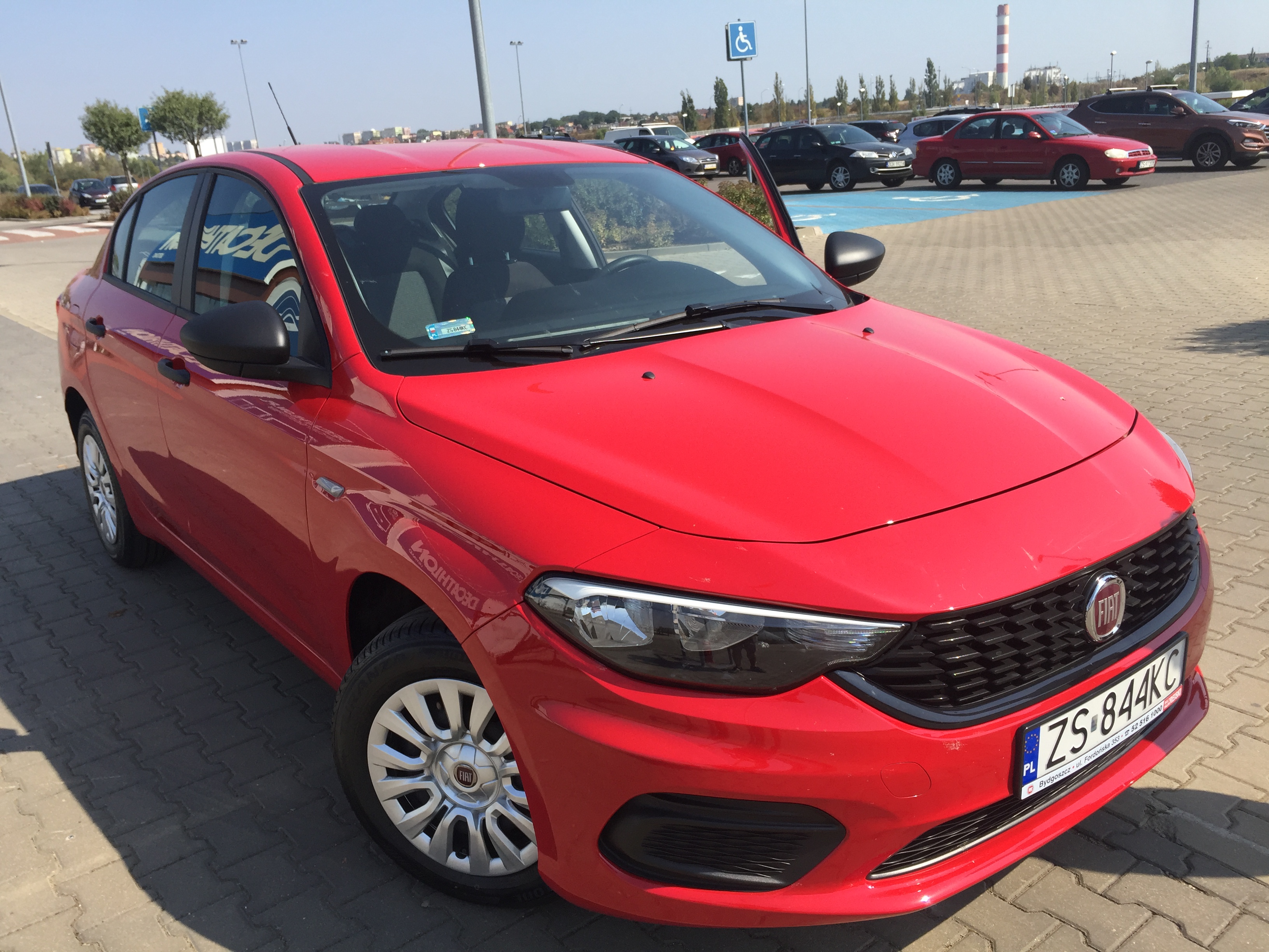 Czerwony samochód osobowy marki Fiat zaparkowany na parkingu z otwartymi drzwiami, widoczne inne pojazdy i znak dla niepełnosprawnych w tle.