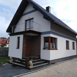 Dwukondygnacyjny dom z białymi ścianami, ciemną dachówką i balkonem z metalową balustradą, z dekoracyjnymi figurami lwa przy wejściu.