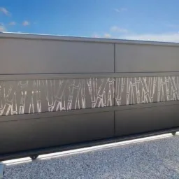 Nowoczesna brama przesuwna w kolorze grafitowym z ażurowym, geometrycznym wzorem na tle błękitnego nieba. Widoczne słupki i kamienny podjazd.