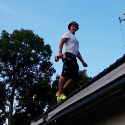 Dekarz z wiertarką akumulatorową na dachu, w kapeluszu i neonowych butach, na tle błękitnego nieba i zielonych drzew.