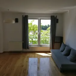 Jasne wnętrze pokoju z drewnianą podłogą, grzejnikiem pod oknem z widokiem na zieleń, szarą sofą z poduszkami i nowoczesnym oświetleniem.