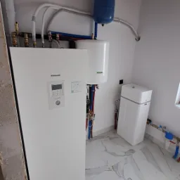 Biała instalacja grzewcza Panasonic Aquarea Smart Cloud z niebieskim zbiornikiem wyrównawczym i zmiękczaczem wody w pomieszczeniu z marmurową podłogą i odsłoniętą konstrukcją dachu.