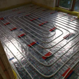 Układanie ogrzewania podłogowego: białe rurki grzewcze zamocowane na folii z czerwoną kratką, widok na okno z odbijającym się światłem słonecznym.