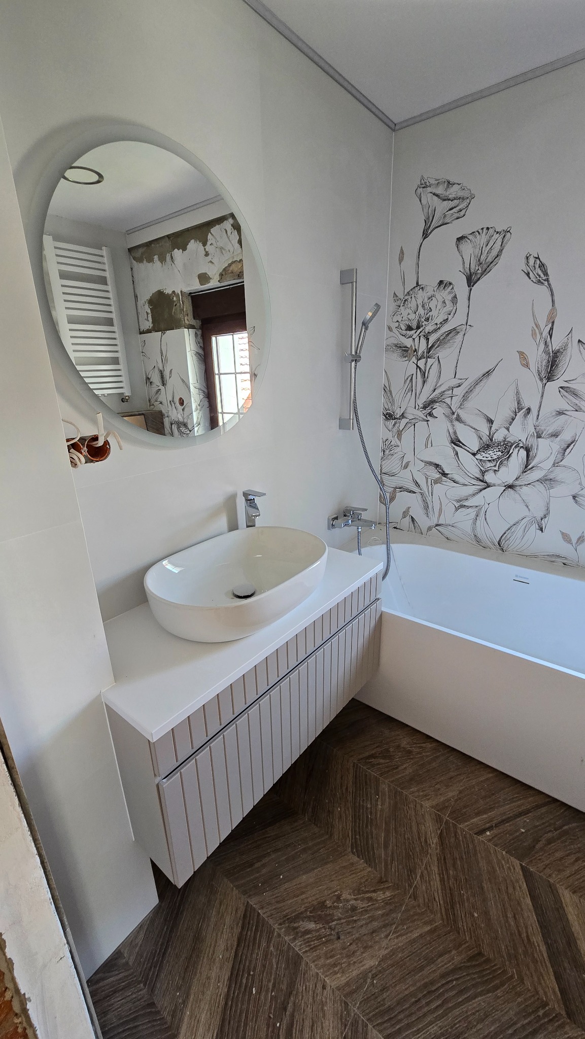 Nowoczesna łazienka po remoncie: biała umywalka nablatowa, szafka z lamelami, wanna i dekoracyjna tapeta w kwiaty. Parkiet ułożony w jodełkę. W lustrze odbicie remontowanego pokoju.