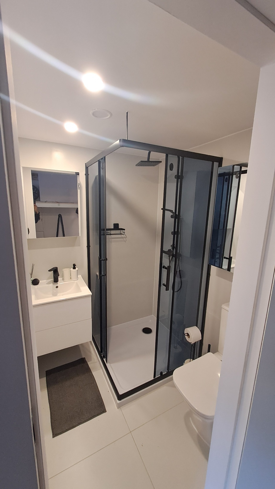 Nowoczesna łazienka po remoncie: kabina prysznicowa typu walk-in z czarnymi ramami, biała umywalka z szafką, lustro z szafką i biała toaleta. Minimalistyczny design, jasne płytki.