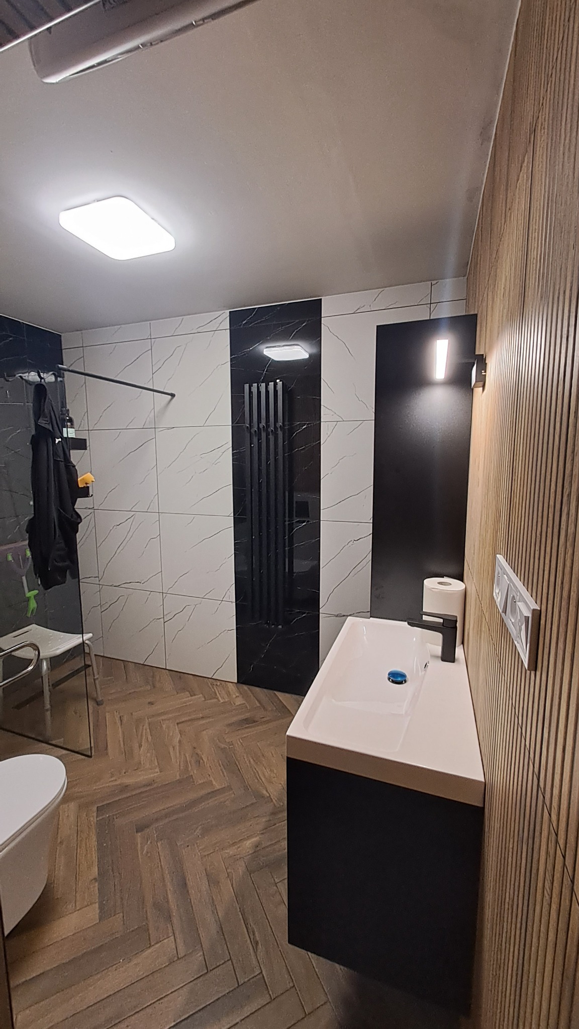 Nowoczesna łazienka po remoncie: biała umywalka z czarną szafką, grzejnik drabinkowy, płytki imitujące marmur i drewnopodobna podłoga w jodełkę.